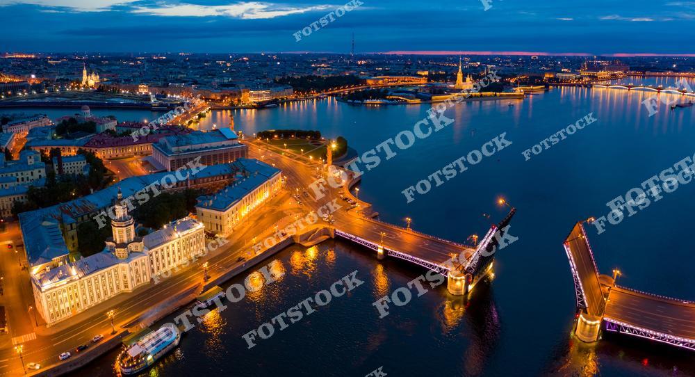 На фотографии изображен разведенный Дворцовый мост, освещенный огнями, с видом на реку Неву и Петропавловскую крепость. Справа от моста виднеется Петропавловская крепость, один из исторических символов Санкт-Петербурга. Слева — здание Кунсткамеры, одного из старейших музеев России, и ансамбль зданий Стрелки Васильевского острова. Ночное освещение и отражение огней в воде придают изображению особую атмосферу.